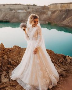 a woman in a wedding dress is standing on rocks near the water and holding a bouquet