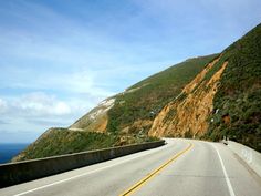 an empty highway with the ocean in the back ground and hills on either side that are very steep