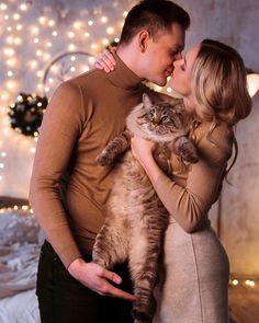 a man and woman kissing while holding a cat in front of a bed with christmas lights