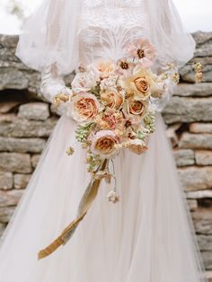 a bridal holding a bouquet of flowers in her hand and standing next to a stone wall