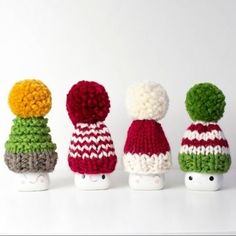 four small knitted toys are lined up in a row on a white surface, one is wearing a striped hat and the other has a multi - colored pom