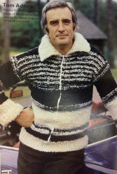 an older man wearing a sweater and jacket in front of a car with his hands on his hips