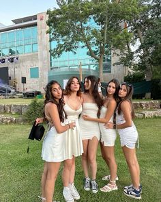 four women in white dresses posing for a photo together on the grass near a building