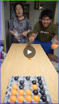 two people standing at a table with oranges in front of them and an egg carton on the table