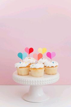 cupcakes on a cake stand with colorful paper heart picks in the middle, against a pink background