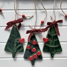 three ceramic christmas tree ornaments hanging from a rope on a white wall with red berries and twine