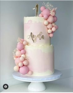 a pink and white two tiered cake with balloons on the top that spell it's first birthday