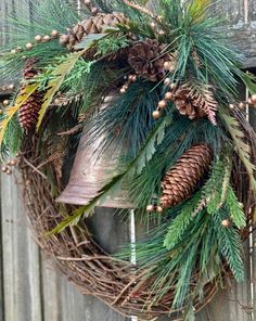 a wreath with pine cones and evergreens hanging on a wooden fence next to a bell