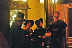 four people dressed in old fashioned clothing stand outside a building at night, one man is holding an open book and the other woman is wearing a top hat