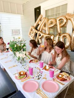 three women sitting at a table with pink plates and gold foil balloons in the background