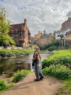 a woman standing on a path next to a river with buildings in the back ground