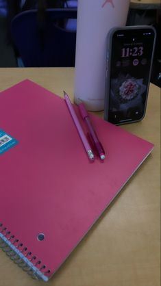 a pink notebook, cell phone and pen on a table in front of a cup