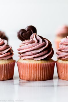 three cupcakes with pink frosting and chocolate hearts on top, sitting in front of each other