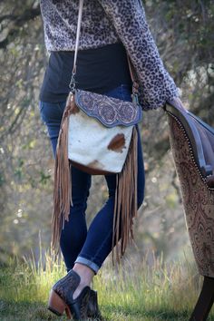 "Presenting the Cactus Blossom Hand-Tooled Fringe Bag.  The leather flap on this bag is carved by an intricate pattern of flowers.  The body of the purse is brown and white cowhide and it has fringes on both sides. Pair it with a boho dress and add a spark to your look!  It is a roomy cross body style purse, so you can use it everyday!  The cowhides vary  Some of the bags I have are more of the brown or tan hides.  The picture shows a bag with more white.  Please send a note to let me know what Myra Bags Cowhide, Western Style Bags With Concho, Western Bags With Concho Detail, Hand Tooled Western Bags, Brown Hand Tooled Bag For Rodeo, Hand Tooled Brown Bag For Rodeo, Hand Tooled Brown Bags For Rodeo, Hand Tooled Leather Bags For Rodeo, Western Style Crossbody Bags For Western-themed Events