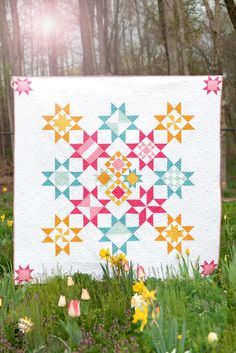 a colorful quilt sitting on top of a lush green field next to trees and flowers