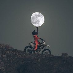 a person on a motorcycle holding the moon up in the air above their head while standing on top of a hill