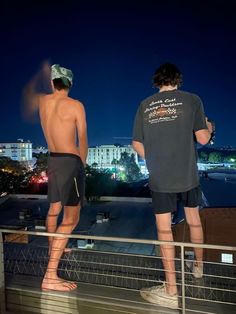 two men standing on top of a roof looking at the city lights in the distance