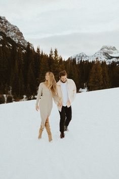 an engaged couple walking through the snow in front of some trees and mountains at sunset