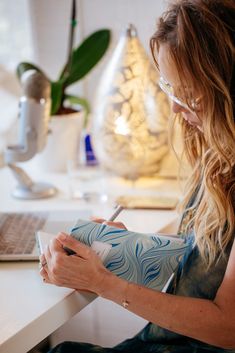 a woman sitting at a desk writing on a piece of paper with a laptop in the background