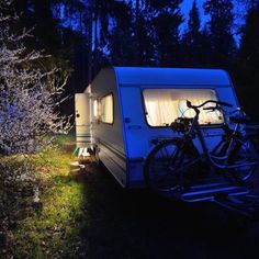 a camper trailer parked in the woods at night with its lights on and two bicycles attached to it