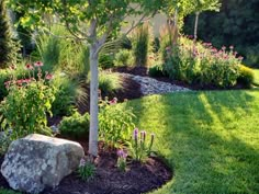 a rock in the middle of a garden with trees and flowers around it, surrounded by grass