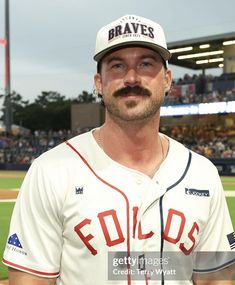 a man with a mustache standing in front of a baseball field wearing a white and red uniform