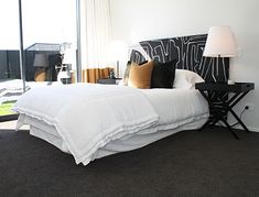 a bed with white sheets and pillows next to a night stand in front of a large window