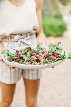 a woman holding a plate with salad on it in her hands and wearing shorts around her waist