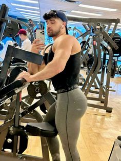 a man standing in front of a gym machine looking at his cell phone while wearing leggings