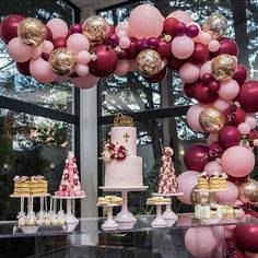 a table filled with cakes and desserts under a large balloon arch in front of a window