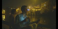 two people sitting on a bench in front of the water at night with lights reflecting off them