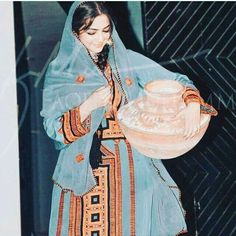 a woman wearing a blue and orange dress holding a bowl in her hand while standing next to a wall