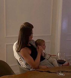 a woman holding a child while sitting at a table with a glass of wine in front of her