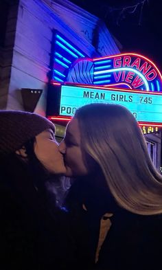 two women are kissing in front of a theater sign at night with neon lights on the marquee