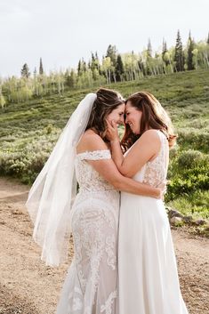 two brides hugging each other in front of a field with trees on the hill