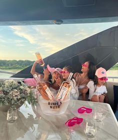 three women sitting at a table with drinks and sunglasses on their heads, one holding up a bottle