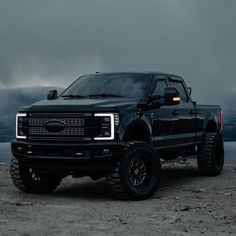 a large black truck parked on top of a rocky hill next to the ocean with mountains in the background