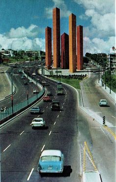 cars driving down the road in front of tall red buildings with orange towers on top