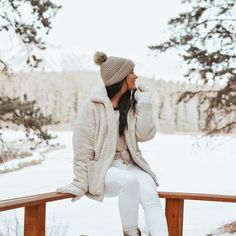 a woman sitting on top of a wooden bench next to snow covered ground and trees