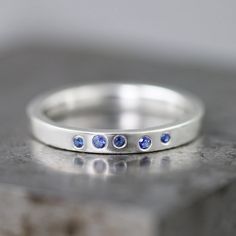 "This sterling silver ring can be worn as an alternative engagement ring, a wedding band, or just a stunning everyday ring to stack with any other rings in your collection. Five small flush set gorgeous blue sapphires in two sizes are scattered along one side of the ring. The pattern of the stones in the ring I make for you might be slightly different than the pattern shown in the photos, as each ring is made one by one to order, but the ring will look very similar. This band measures 2.5mm wide Wedding Sapphire Ring In Sterling Silver, Modern Stackable Wedding Rings With Birthstone, Modern Stackable Sapphire Wedding Ring, Wedding Sapphire Ring With Polished Sterling Silver, Blue Stackable Wedding Rings With Bezel Setting, Wedding Sapphire Stackable Ring In Sterling Silver, Wedding Sapphire Ring In Sterling Silver With Polished Finish, Silver Sapphire Stackable Rings For Wedding, Minimalist Silver Sapphire Ring With Round Band