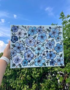 a hand holding up a blue and white tile with flowers on it in front of some trees