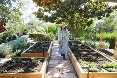 a woman wearing a hat and coat standing in a garden filled with lots of plants