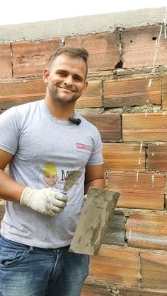 a man standing in front of a brick wall holding a piece of wood and wearing gloves