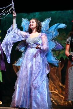 a woman dressed in fairy costume holding an umbrella and smiling at the camera while standing on stage