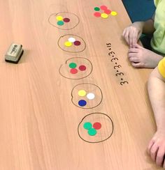 two children are sitting at a table with numbers and circles on it, one child is holding a cell phone