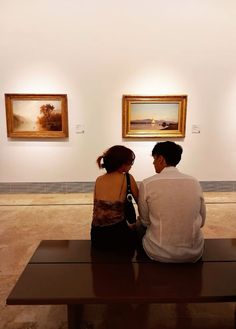 two people sitting on a bench looking at paintings in an art gallery with one person touching the other's ear