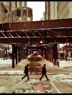 two people crossing the street in front of a train on an overpass with snow