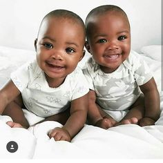 two babies sitting on top of a bed smiling at the camera with white sheets covering them