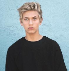 a young man with blonde hair standing in front of a blue wall wearing a black sweater