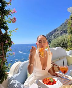 a woman sitting at a table with food and drinks in front of her, overlooking the ocean